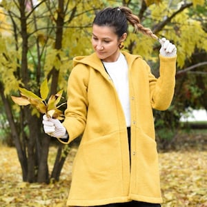 May include: A woman wearing a yellow hooded coat with a white shirt underneath. She is standing in a park with fallen leaves on the ground.