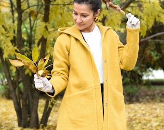 May include: A woman wearing a yellow hooded coat with a white shirt underneath. She is standing in a park with fallen leaves on the ground.