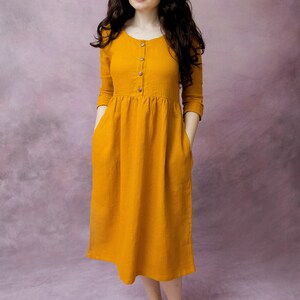 May include: A woman wearing a mustard yellow linen dress with a button-up front and pockets. The dress has a gathered waist and falls to the mid-calf.
