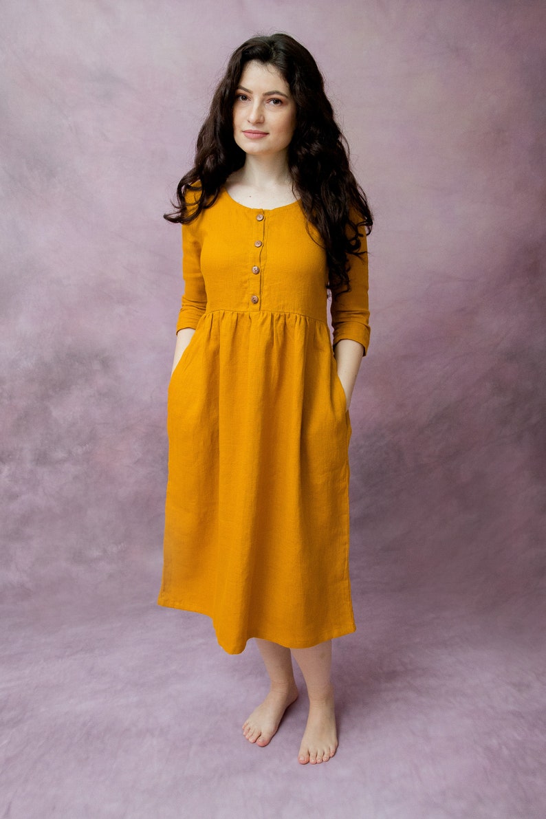May include: A woman wearing a mustard yellow linen dress with a button-up front and pockets. The dress has a gathered waist and falls to the mid-calf.