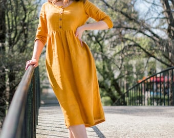 Pode incluir: Uma mulher vestindo um vestido de linho amarelo mostarda com botões na frente e mangas três quartos. Ela está em pé em uma ponte com uma grade e usando sandálias de cunha brancas.