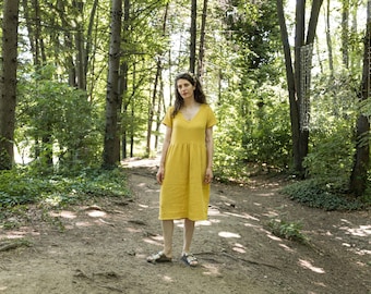 Può includere: Una donna indossa un abito giallo a maniche corte con scollo a V, in piedi su un sentiero nel bosco. L'abito, ampio e lungo fino a metà polpaccio, contrasta con il verde degli alberi. Indossa sandali.