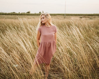 Può includere: Un abito color rosa antico con maniche corte e vita arricciata. L'abito, indossato da una persona, arriva al ginocchio e si trova in un campo di erba alta e secca. Lo sfondo presenta un paesaggio naturale e morbido.
