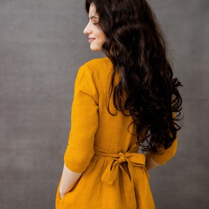 May include: A woman wearing a mustard yellow linen dress with a tie at the waist. The dress has pockets on the sides.
