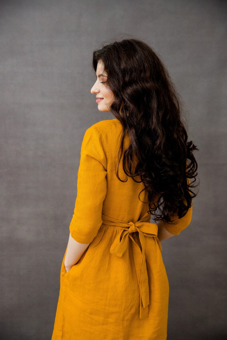 May include: A woman wearing a mustard yellow linen dress with a tie at the waist. The dress has pockets on the sides.