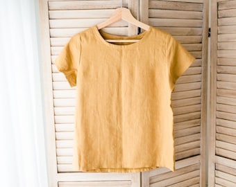 May include: A mustard yellow linen t-shirt with short sleeves hanging on a wooden hanger.