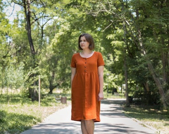 Può includere: Una donna con un vestito di lino color ruggine cammina su un sentiero in un parco. Il vestito ha una chiusura con bottoni sul davanti e maniche corte.