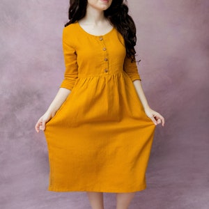 May include: A woman wearing a long, mustard yellow, linen dress with a button-up front and 3/4 sleeves. She is standing on a purple background.