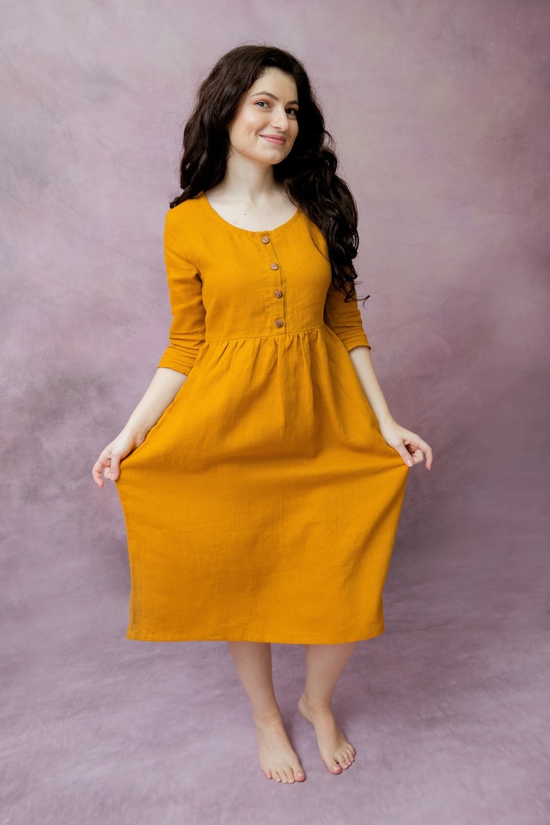 May include: A woman wearing a long, mustard yellow, linen dress with a button-up front and 3/4 sleeves. She is standing on a purple background.