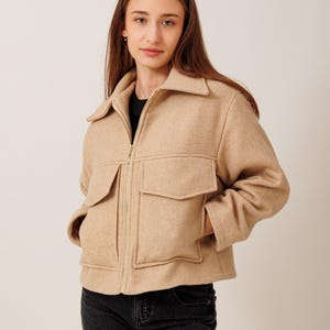 May include: A tan-colored jacket with a collared neckline, two front pockets, and a gold zipper. The jacket has long sleeves and a slightly cropped fit. The model is wearing a black shirt underneath and black jeans.