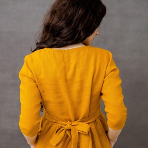 May include: A woman wearing a long, mustard yellow dress with a tied sash at the waist. The dress has long sleeves and pockets on the sides.