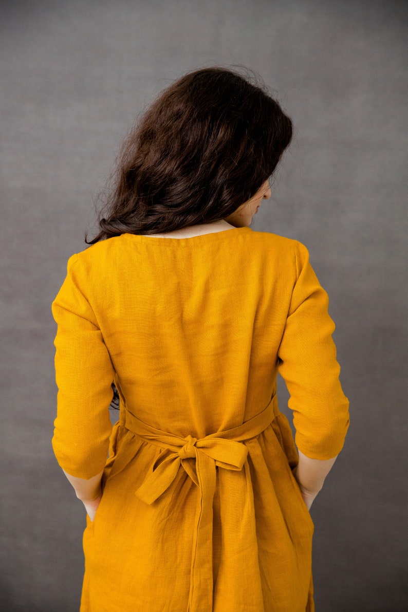 May include: A woman wearing a long, mustard yellow dress with a tied sash at the waist. The dress has long sleeves and pockets on the sides.