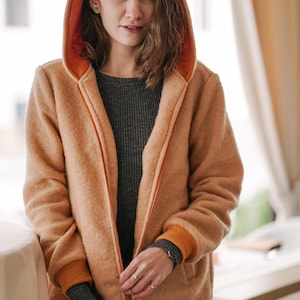 Op de afbeelding: Een bruinbeige jas met capuchon en een oranje binnenkant en bies. De jas heeft een ritssluiting en geribde manchetten met oranje accenten. De persoon draagt er een grijze trui jurk onder.