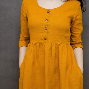 May include: A mustard yellow linen dress with a round neckline and a button-up front. The dress has long sleeves and a gathered skirt.