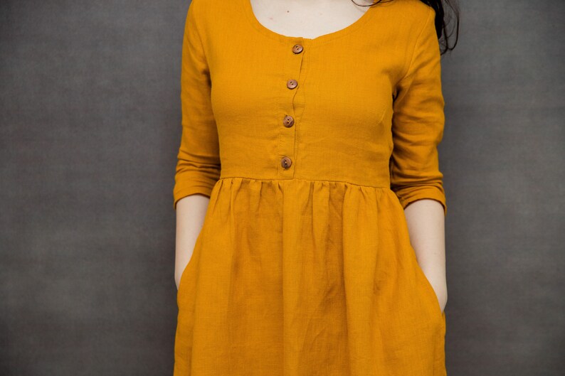 May include: A mustard yellow linen dress with a round neckline and a button-up front. The dress has long sleeves and a gathered skirt.