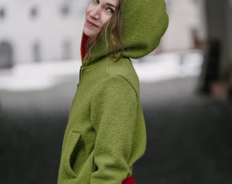 May include: A woman wearing a green hooded jacket with red trim. The jacket has a zipper closure and a pocket on the front. The woman is standing in front of a white building.