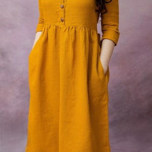 May include: A mustard yellow linen dress with a round neckline, three-quarter sleeves, and a button-up front. The dress has two pockets and a gathered waist.