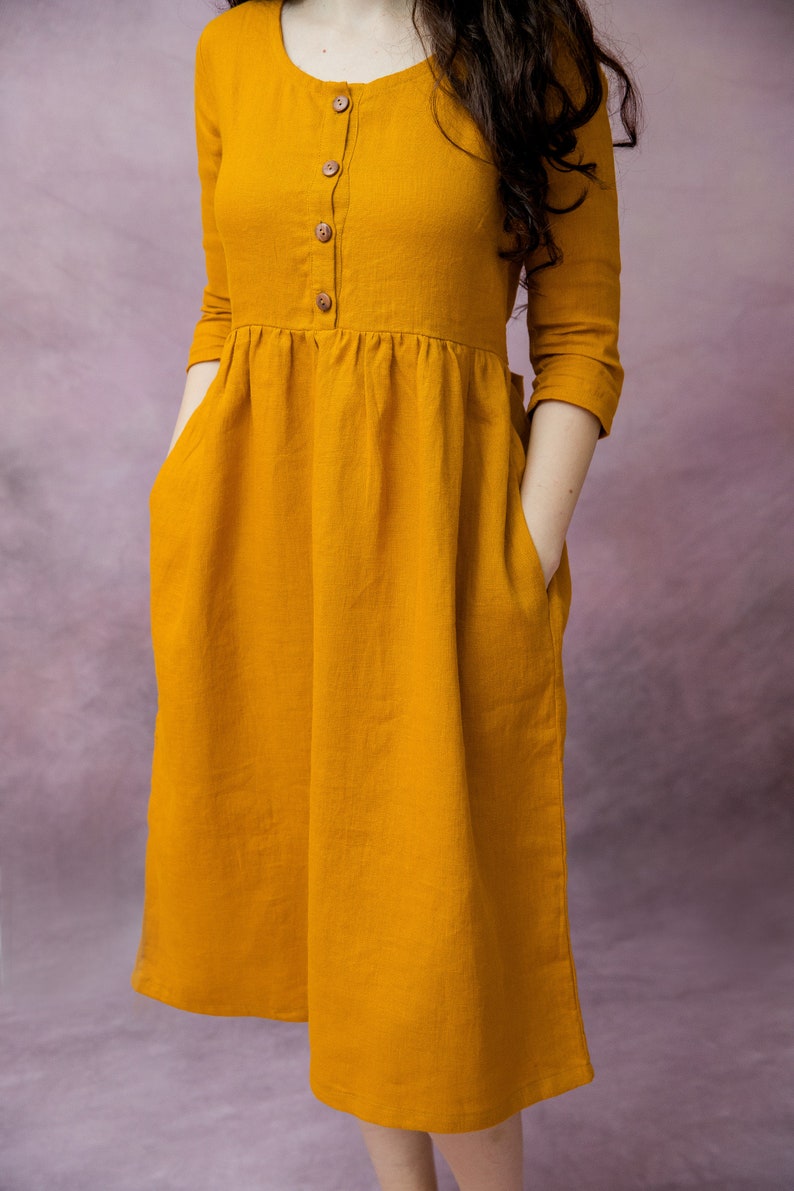 May include: A mustard yellow linen dress with a round neckline, three-quarter sleeves, and a button-up front. The dress has two pockets and a gathered waist.