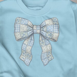 May include: A pale blue sweatshirt with a large bow design. The bow is a patchwork of light blue and white fabrics with floral and polka dot patterns. The sweatshirt is made of a soft material.