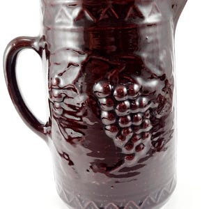 May include: A dark brown glazed ceramic pitcher featuring a raised relief design of grape clusters and leaves on the front. The vessel has a sturdy handle, a pouring spout, and decorative geometric triangle patterns along the top and bottom rims.