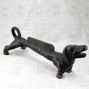 May include: A dark, weathered cast iron boot scraper shaped like a dachshund dog. The elongated body features a vertical blade along the back for cleaning footwear, with the dog's head facing forward and its tail curled upward in a loop.