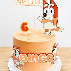 Op de afbeelding: Een oranje cake met cartoonkarakteruitsparingen en de naam "Clariss". De taart heeft een "6" kaars en het woord "Bingo" aan de zijkant. De taart staat op een wit bord.