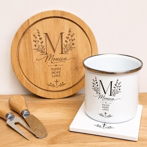 Pode incluir: Uma tábua de corte redonda de madeira, uma caneca de esmalte branco com borda prateada, duas facas de queijo de metal com cabos de madeira e um porta-copos quadrado branco. Cada item apresenta a letra M, o nome Monica e o texto FAITH HOPE LOVE.