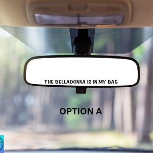 May include: A rearview mirror in a car with the text "THE BELLADONNA IS IN MY BAG" printed on it. The mirror is reflecting a blurry image of trees outside the car window. The text "OPTION A" is printed below the mirror.