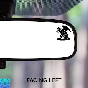 May include: A black gargoyle sticker is affixed to a car's rearview mirror. The sticker is a silhouette of a seated gargoyle with outstretched wings. The text "FACING LEFT" is printed in black below the mirror.