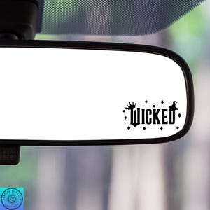 May include: A black and white sticker with the word "Wicked" in a gothic font, adorned with stars, a crown, and a witch hat. The sticker is displayed on a car's rearview mirror.