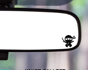 May include: Black ninja decal with a knife, affixed to a car's rearview mirror. The text "KNIFE ON LEFT" is printed below the mirror. The decal is a fun, decorative car accessory.