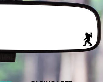 May include: A black Bigfoot silhouette sticker on a car's rearview mirror. The Bigfoot is giving a peace sign. The text "FACING LEFT" is below the mirror.