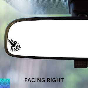 May include: A black silhouette of a hummingbird with its beak in a flower, facing right, is displayed on a white rearview mirror in a car.
