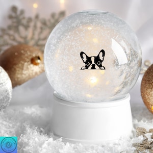 May include: A clear snow globe with a black and white French Bulldog design. The globe sits on a white base and contains white glitter and small lights. Decorative gold and silver ornaments are in the background.
