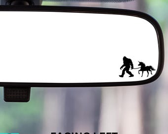 May include: A black and white silhouette of a Bigfoot walking a unicorn, facing left, on a rearview mirror.