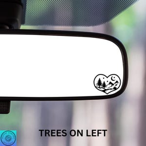 May include: A black heart-shaped decal featuring a mountain scene with trees, stars, and a crescent moon. The decal is placed on a car's rearview mirror. The text "TREES ON LEFT" is at the bottom.