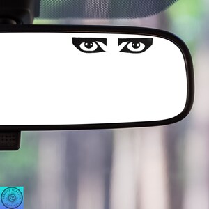 May include: A black and white eye sticker with a dramatic, stylized design. The sticker is placed on a car's rearview mirror.