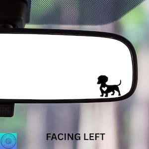 May include: Black dachshund dog silhouette with a heart on a car rearview mirror. The text "FACING LEFT" is below the mirror. The image is set against a blurred background.
