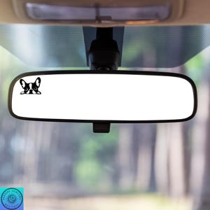 May include: A black and white decal of a French Bulldog peeking over the edge of a car's rearview mirror. The mirror is rectangular with a black frame, mounted inside a vehicle. The background shows a blurred view of trees.