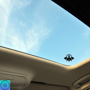 Peut inclure: Une silhouette noire d'un OVNI avec un extraterrestre &agrave; l'int&eacute;rieur, sur un toit ouvrant de voiture contre un ciel bleu avec des nuages blancs.