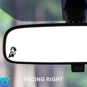 May include: A black and white silhouette of a parrot facing right, placed on the rearview mirror of a car. The mirror is reflecting a blurry image of a green and blue background.