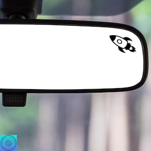 May include: A black and white rocket ship decal on a rearview mirror. The rocket ship is facing to the right and has a small flame coming out of the back.