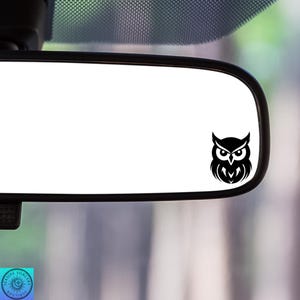 May include: A black owl decal is affixed to the lower right corner of a car's rearview mirror. The owl design is a stylized silhouette with large eyes and a detailed feather pattern. The mirror is rectangular with rounded corners.