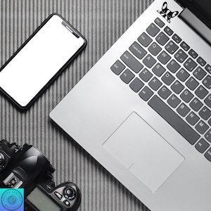 May include: A laptop with a black and white French bulldog sticker on the keyboard, a smartphone, and a digital camera are arranged on a gray textured surface. The laptop is silver, and the phone has a white screen.