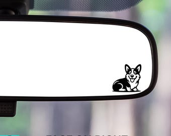 May include: A black and white corgi dog sticker is affixed to a car's rearview mirror. The sticker is a graphic illustration of a corgi dog with its tongue out. The text "FACE ON RIGHT" is below the mirror.