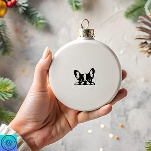 May include: A white ceramic Christmas ornament with a gold-colored metal cap. The ornament features a black and white graphic of a French Bulldog peeking over the edge. The ornament is held in a hand, with a blurred background of greenery.