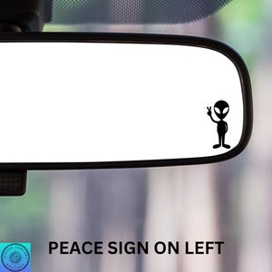May include: A black silhouette of an alien with a peace sign on its left hand, on a white background. The alien is in a car's rearview mirror.