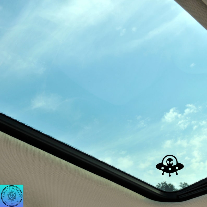 Peut inclure: Une silhouette noire d'un OVNI avec un extraterrestre &agrave; l'int&eacute;rieur, vue &agrave; travers un toit ouvrant d'une voiture contre un ciel bleu avec des nuages blancs.