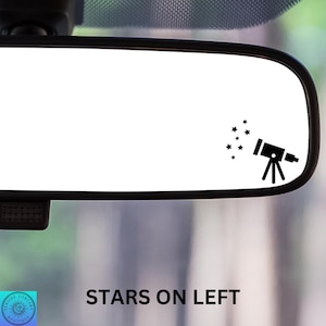 May include: A car rearview mirror with a black frame. The mirror has a black silhouette of a telescope and stars on the right side. The text "STARS ON LEFT" is printed in black at the bottom of the mirror.