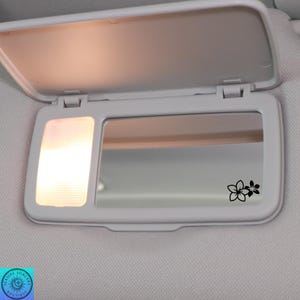 Pu&ograve; includere: Una visiera parasole bianca per auto con specchietto e piccola luce. Lo specchietto presenta un motivo floreale nero. La visiera &egrave; aperta e la luce &egrave; accesa, illuminando l'interno.